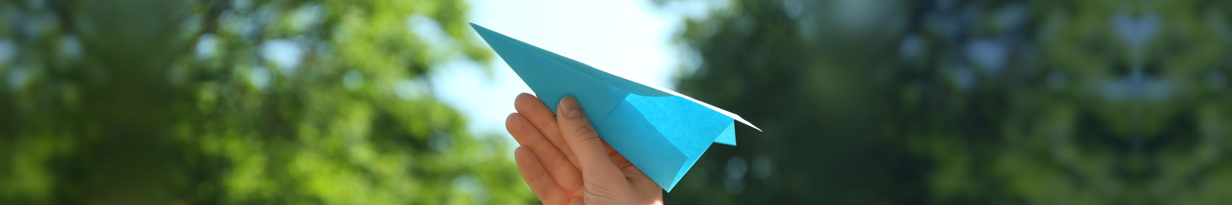 A hand about to launch a blue paper plane against sunlit trees